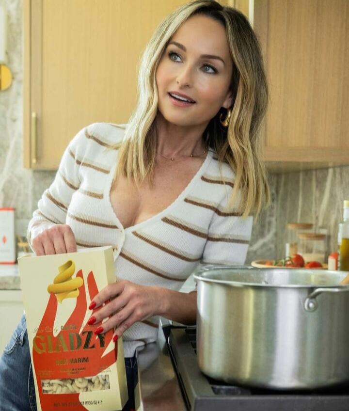 A woman in a striped sweater is in a kitchen, opening a box of pasta next to a pot on the stove.