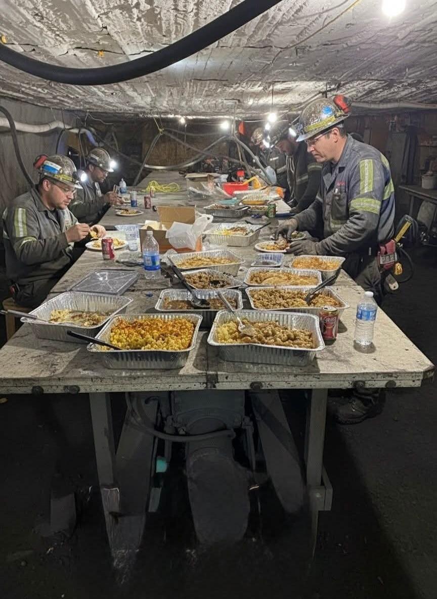 Coal miners seated at a long table underground, eating meals served in aluminum trays. They wear helmets with headlamps and work gear; there are water bottles and canned drinks on the table, and the scene is lit by work lights in a tunnel-like environment.