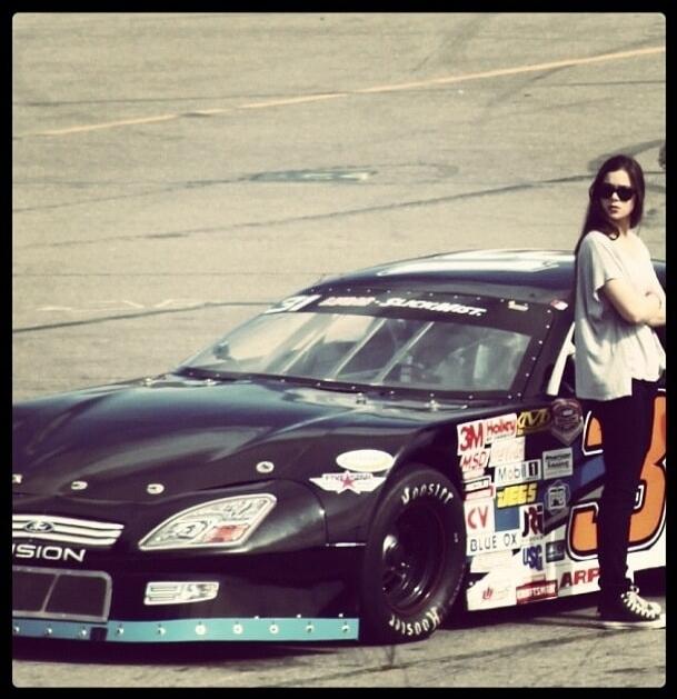 Black race car with sponsor decals parked on a track, a person wearing sunglasses stands beside it.