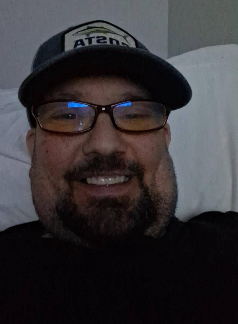 A man wearing a cap with a reversed text 'AT2N' (likely 'JASTA' or 'JUSTA' with a fish logo) and glasses, smiling at the camera.