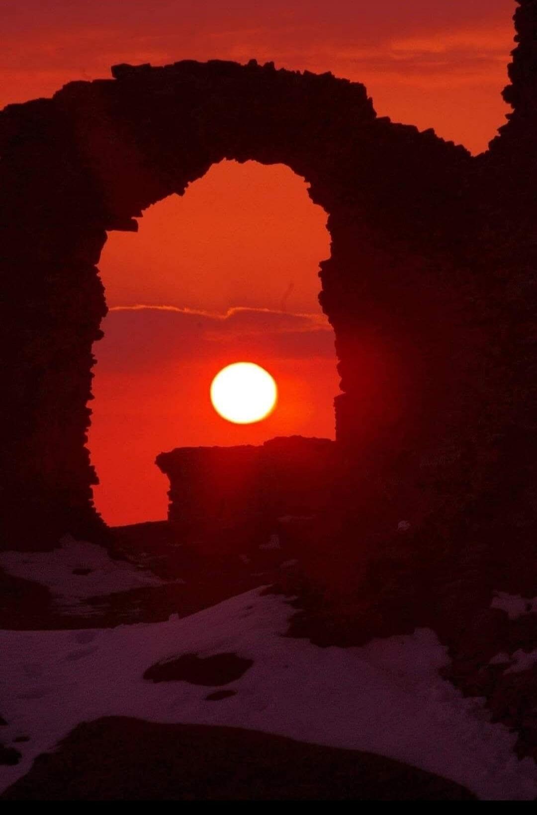 The sun is setting behind a rocky archway, casting a warm, orange glow over the landscape. Snow can be seen on the ground in the foreground.