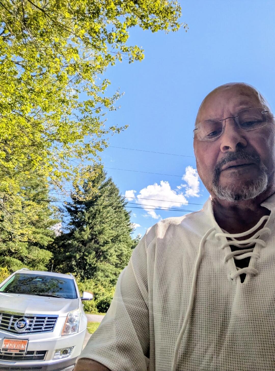 A man takes a selfie outdoors. He has a beard and glasses and is wearing a white waffle-knit shirt. In the background, there are green trees, a blue sky with some clouds, and a silver Cadillac SUV. The license plate on the car reads 'UNIVERSITY OF TENNESSEE' and 'GO VOLS'.