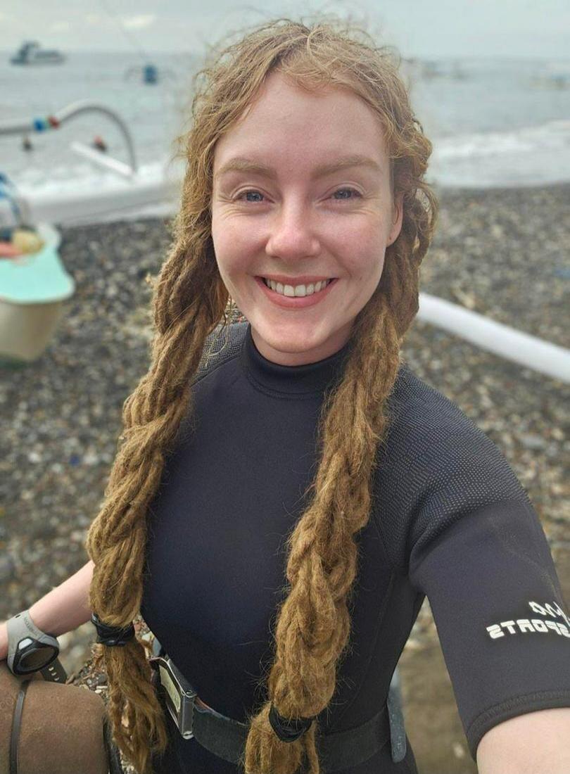 A smiling woman with dreadlocks wearing a black wetsuit stands on a pebbly beach with the sea and a boat in the background. The text on her sleeve appears to be 'ON SPORTS' (mirrored).