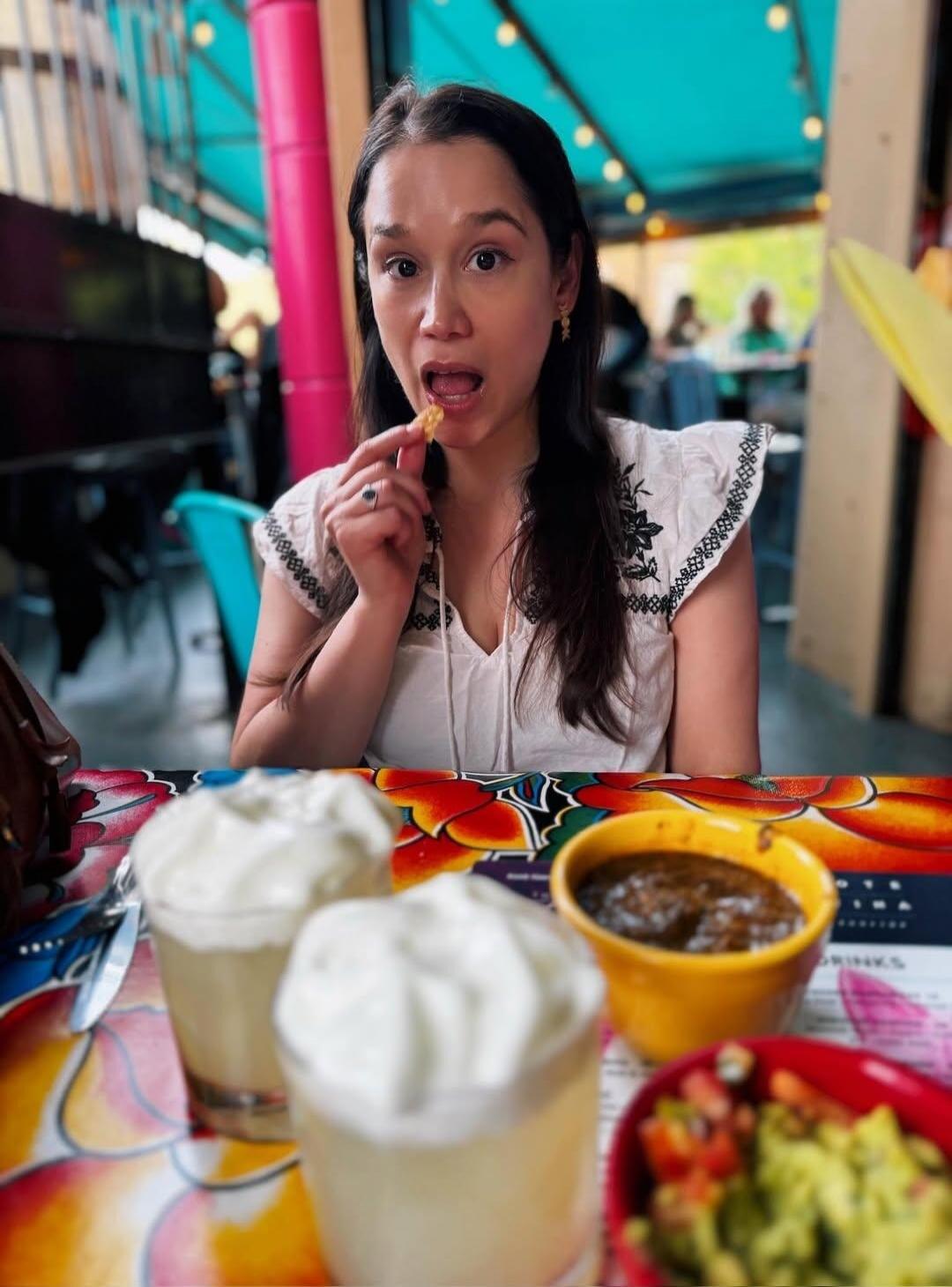 A woman is about to eat a chip at a restaurant table. On the table, there are drinks with whipped cream, salsa, and guacamole.