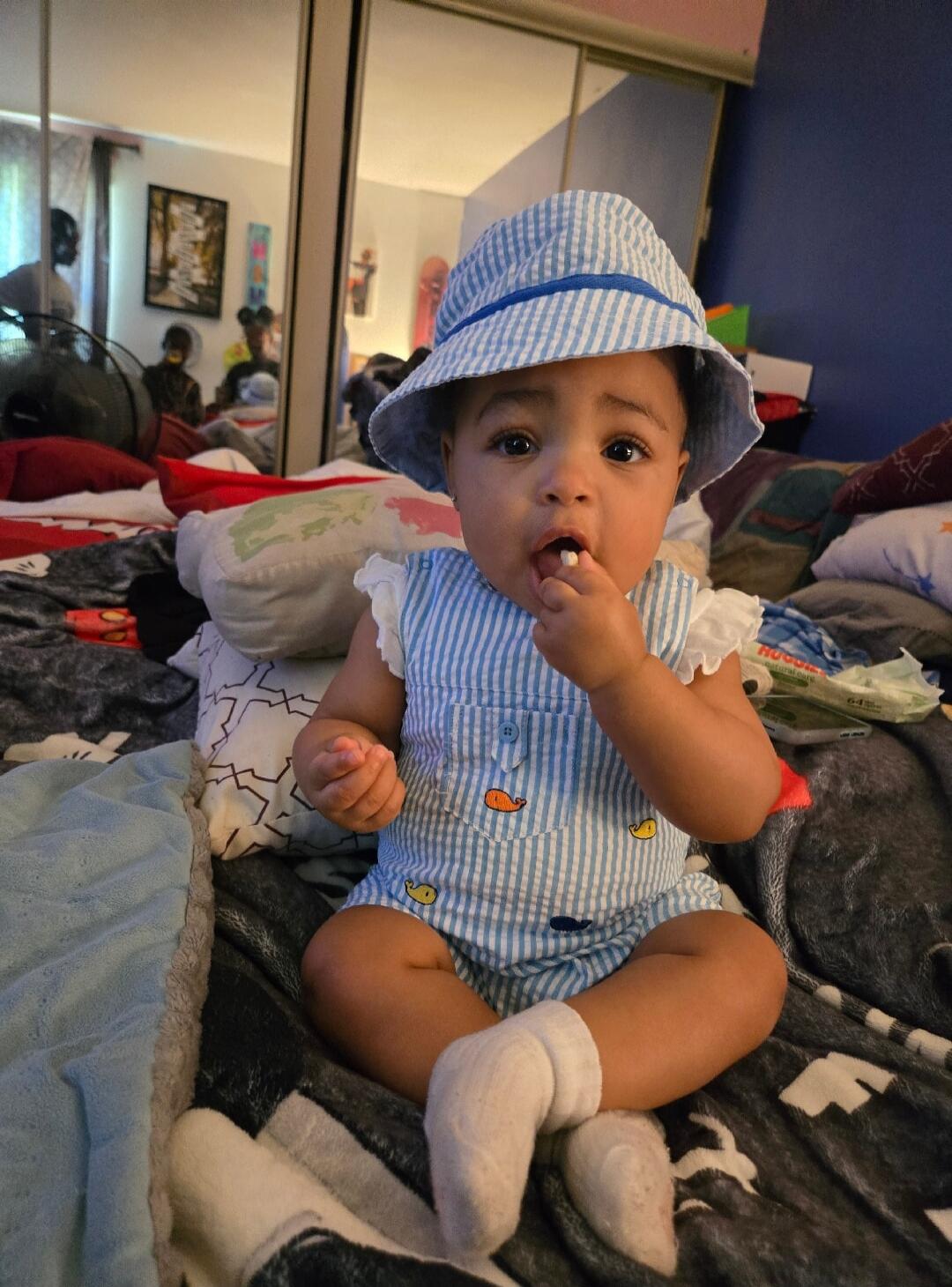 A cute baby wearing a striped hat and romper, sitting on a bed and holding something in its mouth.