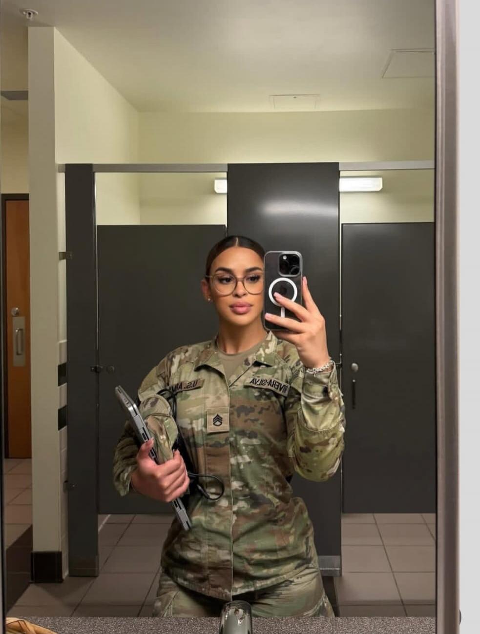 A woman in a military uniform taking a mirror selfie. She is wearing glasses and holding a laptop or tablet. A sergeant rank insignia is visible on her uniform. Text on her uniform appears to be 'MA.20' and 'AVJ2-AIVU' (reversed).