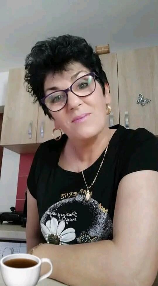 A woman with short dark hair and glasses is looking at the camera. She is wearing a black t-shirt with a floral design and text that appears to be 'EXPERIENCE' (mirrored). She has hoop earrings and a necklace. A cup of coffee is in the foreground. The background shows kitchen cabinets and a butterfly sticker.