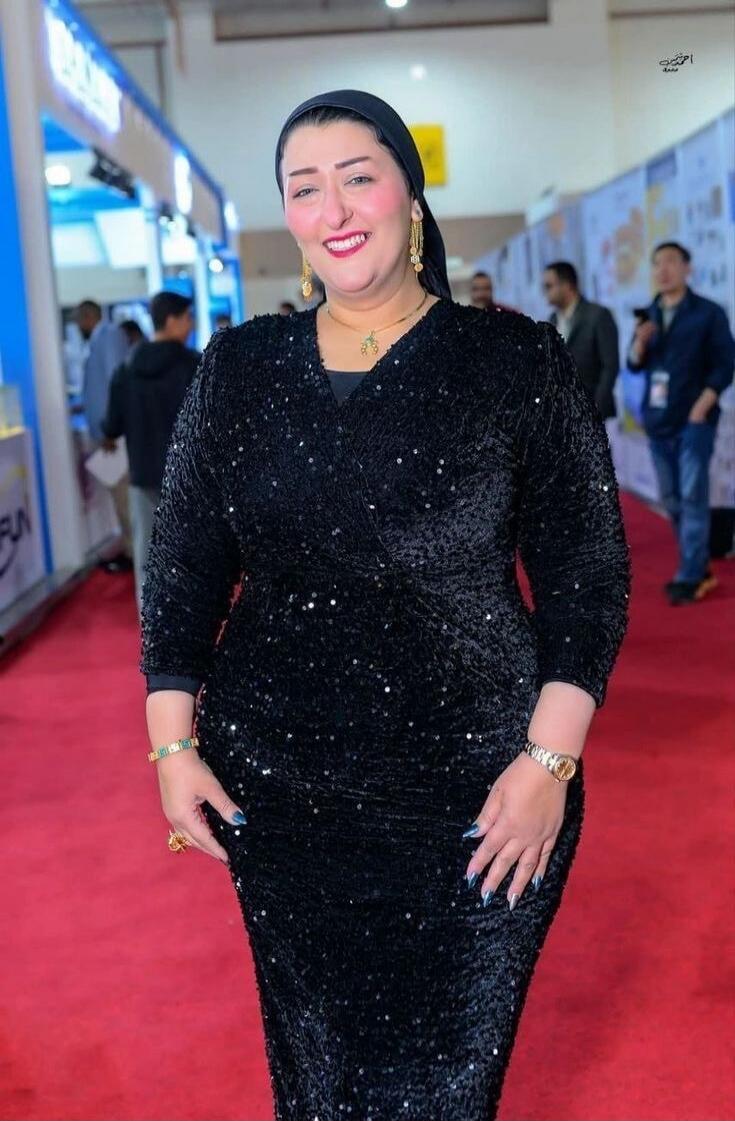 A smiling woman wearing a black sequined dress and a black headscarf poses on a red carpet at an event. She is adorned with gold jewelry. In the background, there are blurred figures and exhibition booths.