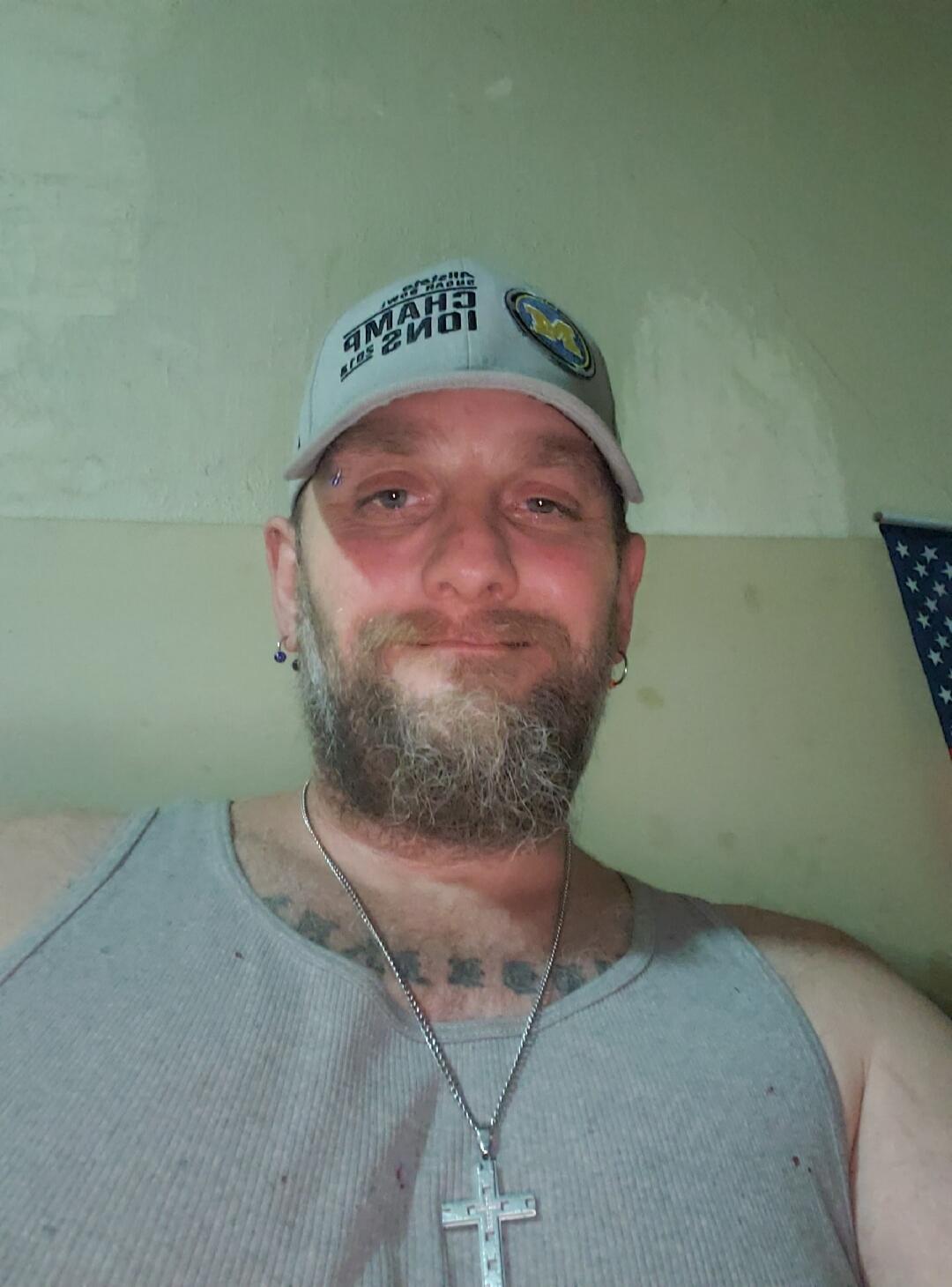 A man wearing a grey tank top, a grey baseball cap with a mirrored text 'CHAMPIONS' and an 'M' logo, and a cross necklace. He has a beard and piercings. A part of an American flag is visible in the background.