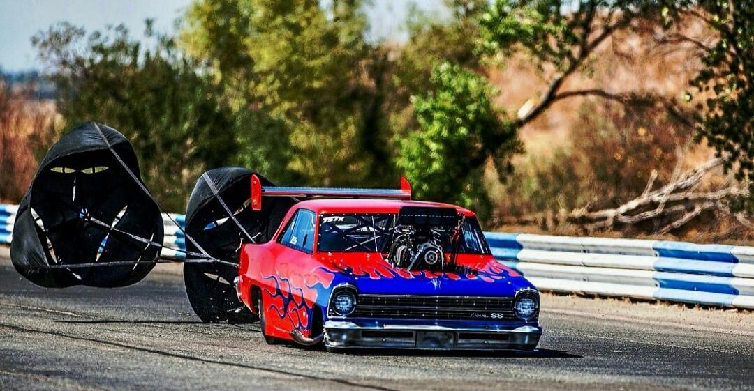 A red and blue drag racing car with flame decals, an exposed engine, and a large spoiler, deploying two drag parachutes to slow down on an asphalt track. The car has '757X' on the window and 'SS' on the grille.
