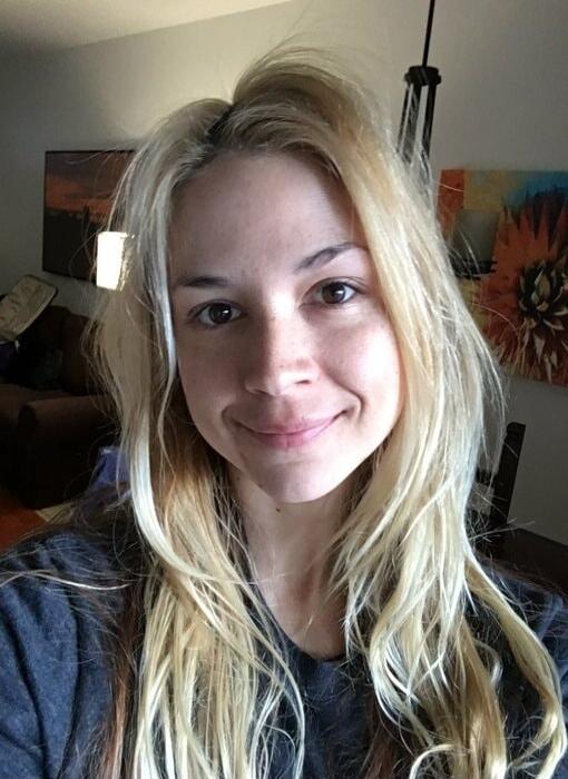 A young woman with long blonde hair taking a selfie indoors, wearing a casual dark shirt, smiling softly at the camera. The background shows living room decor with wall art.