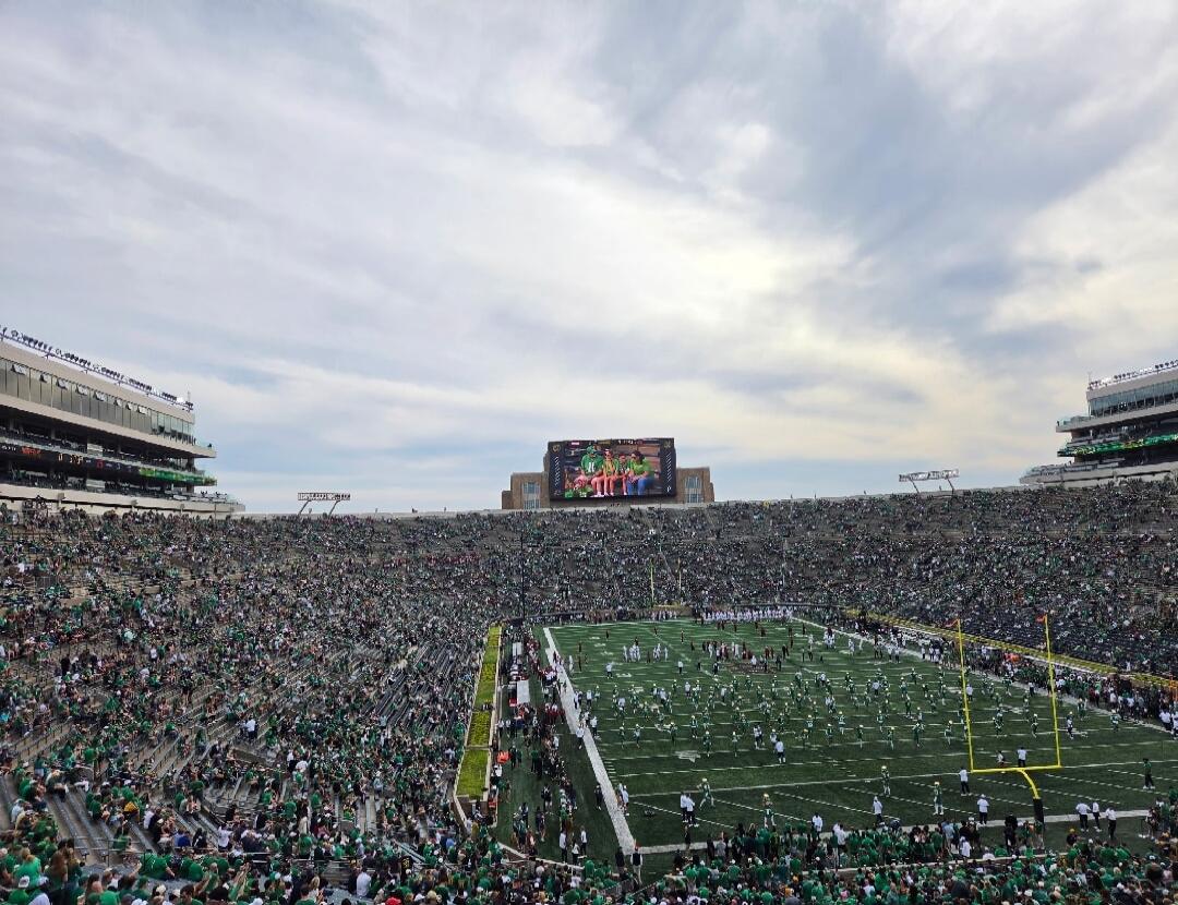 A large American football stadium filled with a crowd, a green field with performers, and a big screen showing a group of people.