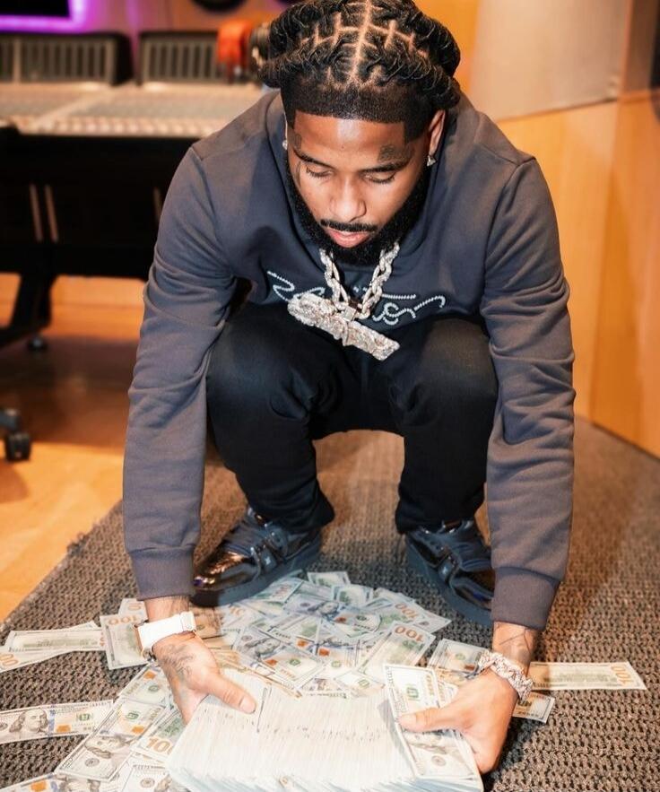 A man with dreadlocks and a diamond chain is squatting over a large pile of US dollar bills, holding a thick stack of money in his hands. The bills are mostly $100 and $10 denominations. He is in what appears to be a recording studio.