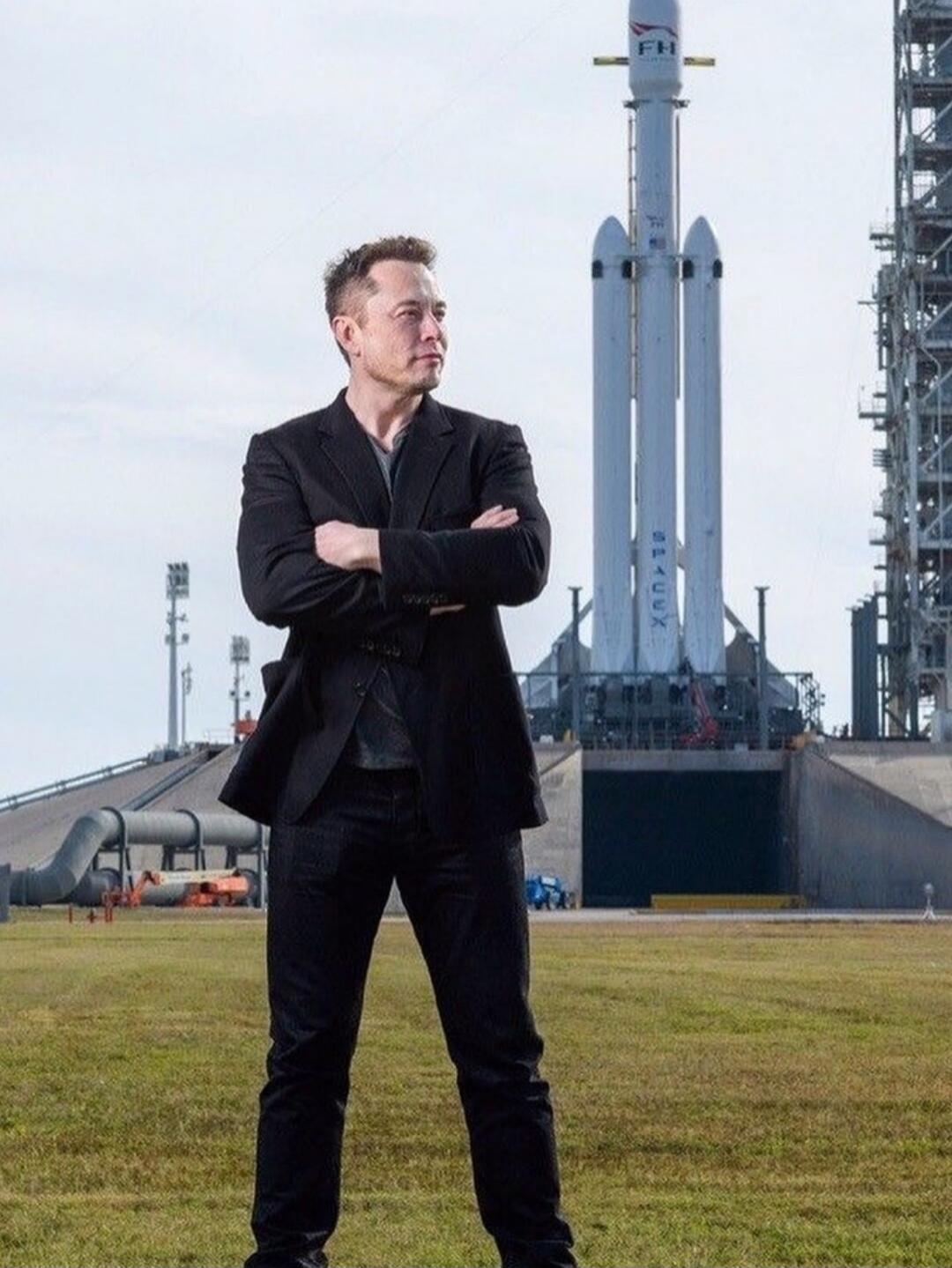 Elon Musk stands in front of a SpaceX Falcon Heavy rocket on a launchpad. The rocket has 'FH' and 'SPACEX' written on it.