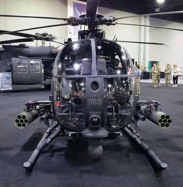A military helicopter, likely an AH-6 Little Bird, with weapon pods mounted on its sides, displayed at an exhibition. The number '369' is visible on its front. Another larger helicopter is in the background, and people, some in military uniform, are observing the display.