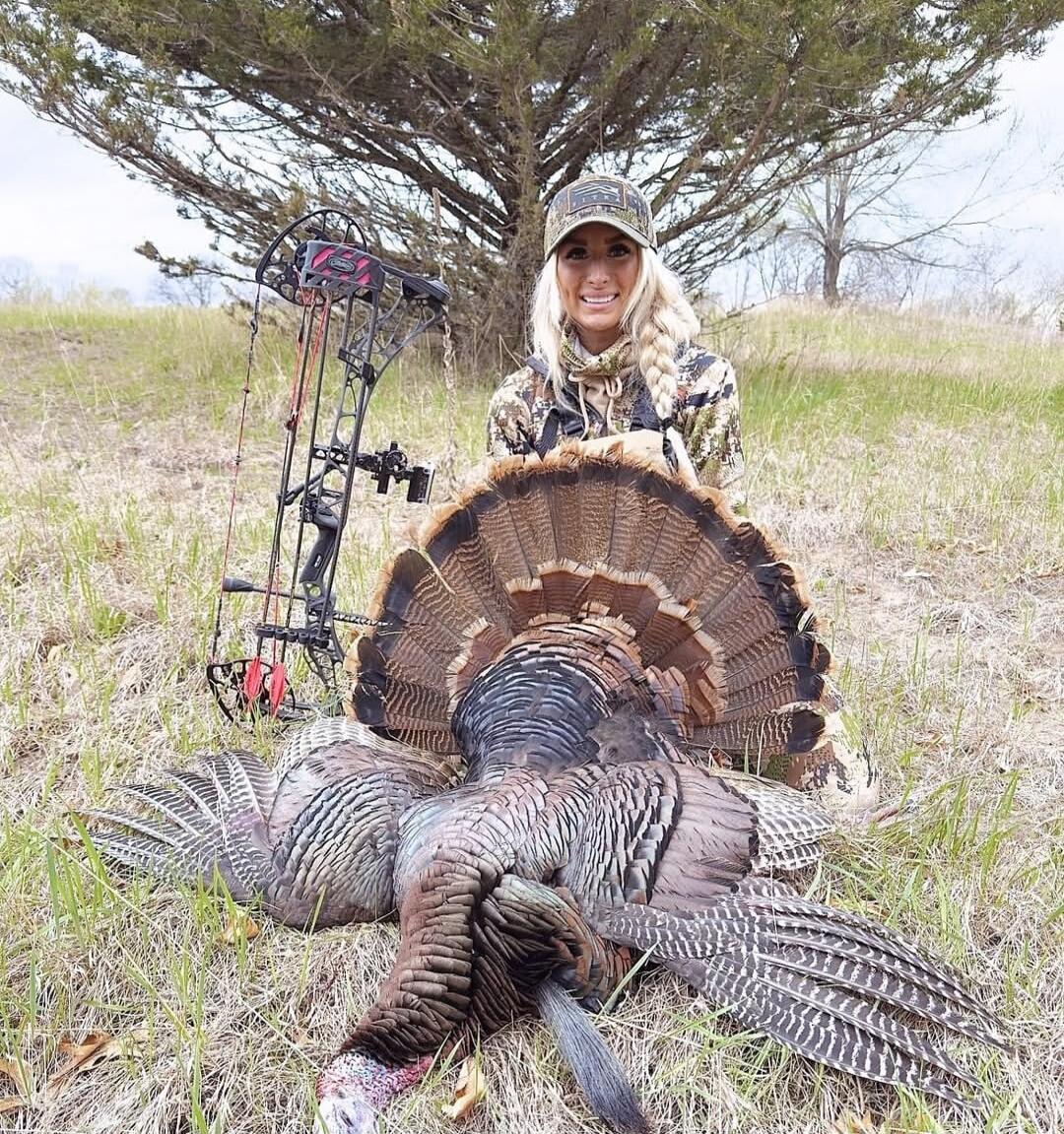 A person in hunting gear kneels next to a large dead wild turkey in a grassy field with a bow in the background.