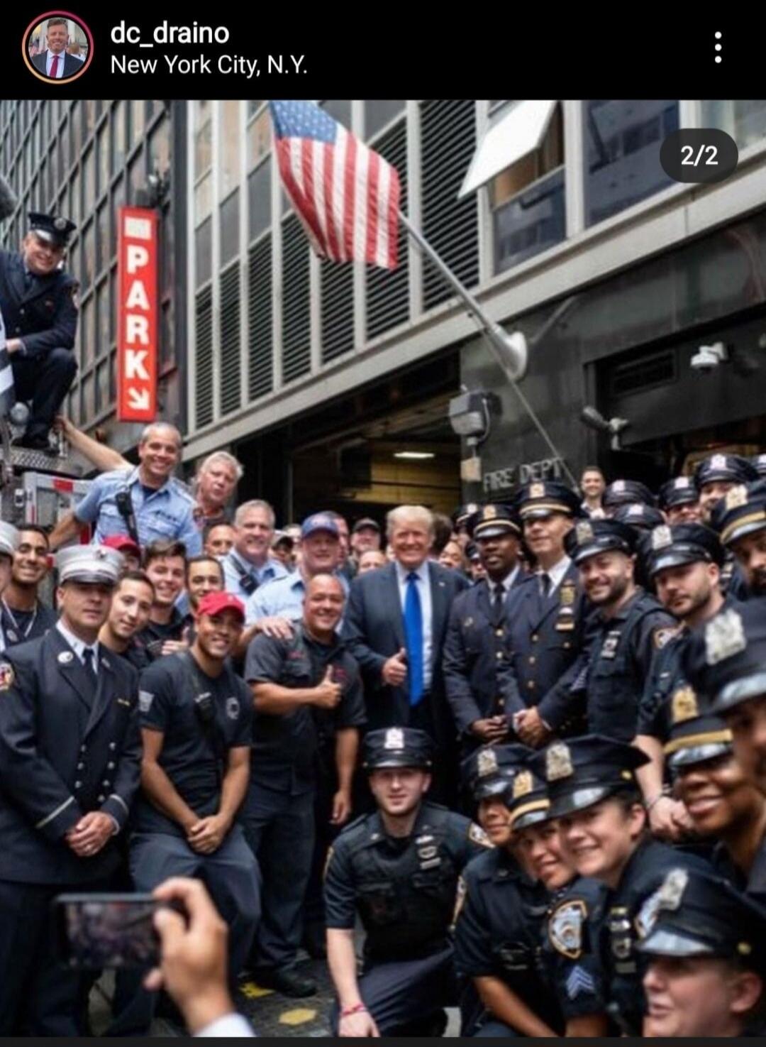 dc_draino New York City, N.Y. PARK FIRE DEPT 2/2. Former President Donald Trump is seen giving a thumbs-up while surrounded by a large group of smiling police officers and firefighters in front of a building with an American flag. Many individuals are taking photos.