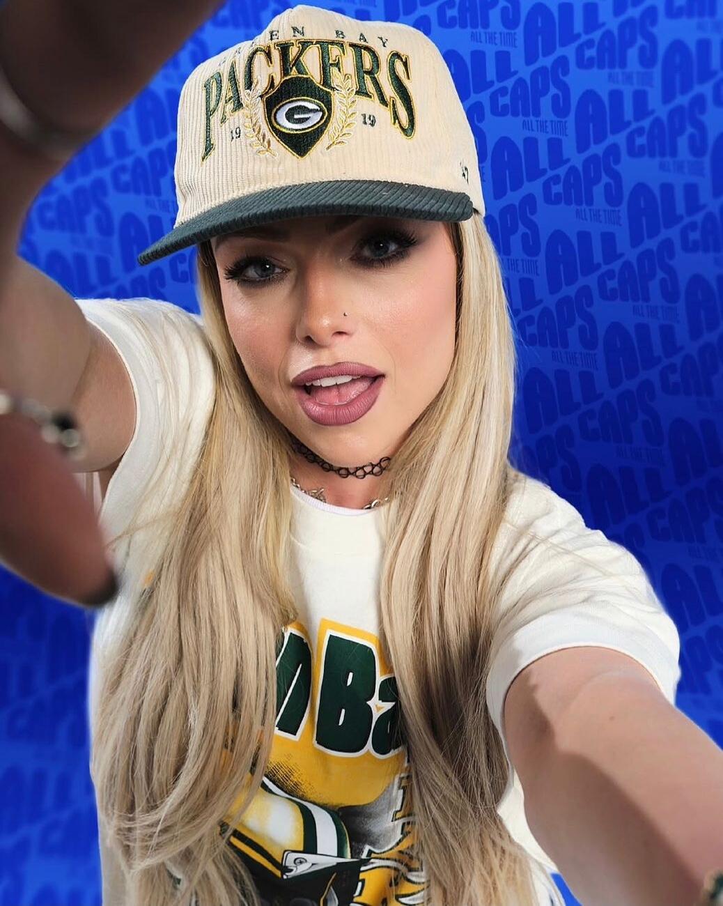 PACKERS on the cap; background with all-caps blue pattern. The person is wearing a white T-shirt and a blonde wig.