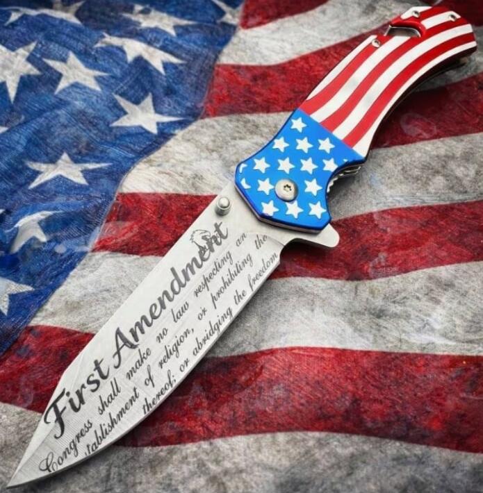A folding knife with an American flag design on its handle, lying on an American flag background. The blade is engraved with text from the First Amendment of the U.S. Constitution: 