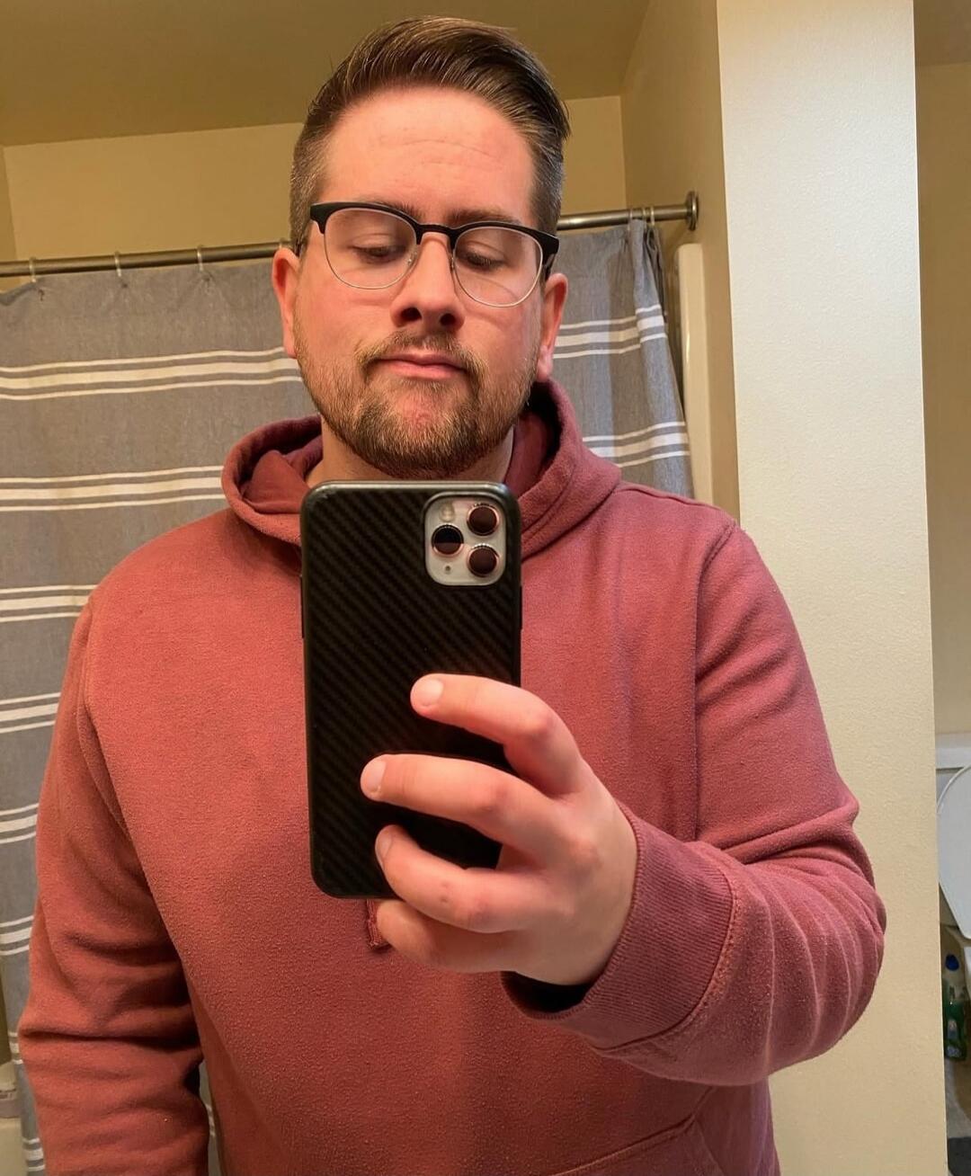 A man in a pink hoodie wearing glasses taking a mirror selfie in a bathroom.