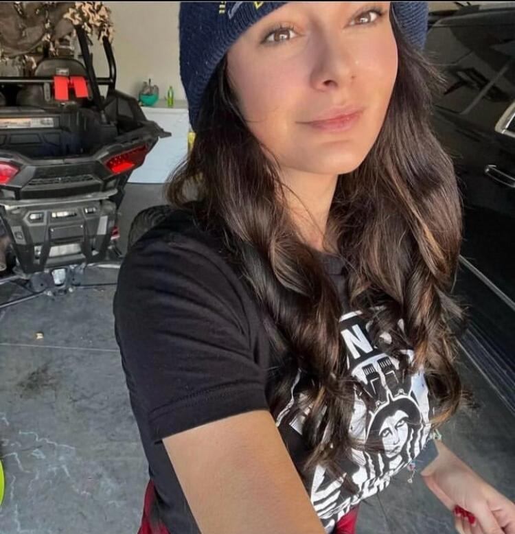 A woman wearing a beanie and a t-shirt, smiling at the camera. An off-road vehicle is visible in the background.