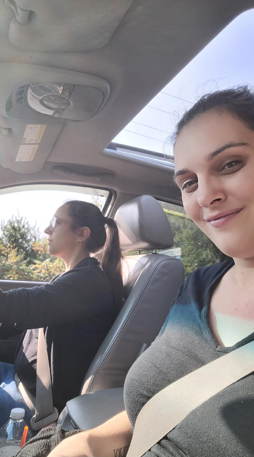 Two women are in a car. One is driving, wearing sunglasses, and the other is in the passenger seat, taking a selfie and smiling. The sun is shining through the sunroof.