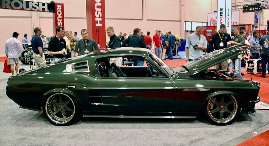ROUSH PERFORMANCE PARTS SEIBON. A dark green classic Ford Mustang with custom wheels and an open hood is displayed at an indoor car show, with several men observing it.