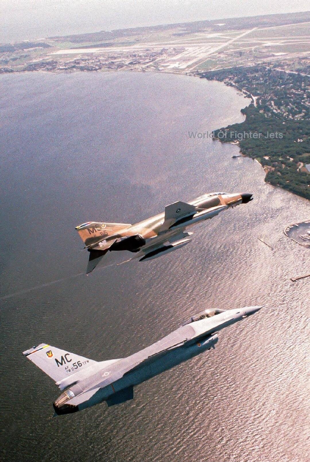 World Of Fighter Jets (watermark)\nTwo fighter jets flying low over a coastline, one camouflaged and one gray, leaving vapor trails across the water.