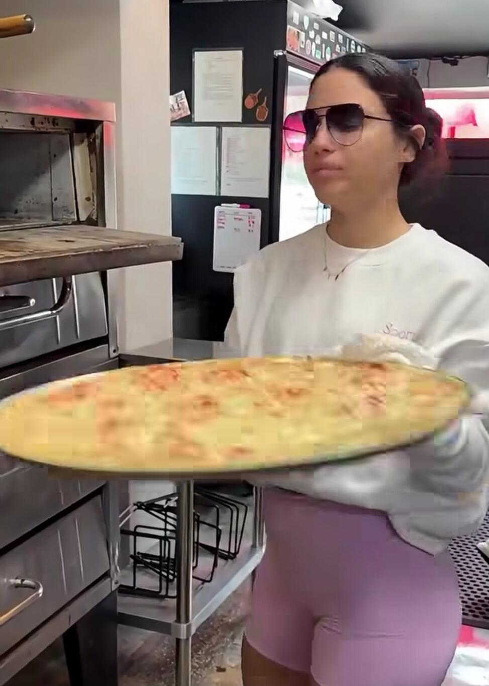 A woman in sunglasses and a white sweatshirt is holding a large cooked pizza on a tray, standing next to a pizza oven in a kitchen.