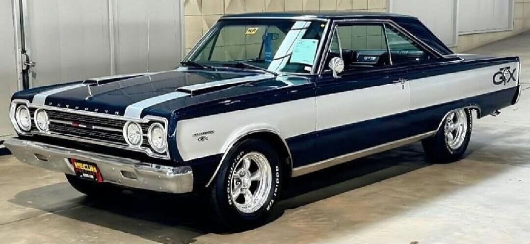 PLYMOUTH GTX MECUM BFGoodrich Radial T/A. A classic American muscle car, the Plymouth GTX, is shown in a two-tone dark blue and silver paint scheme with racing stripes on the hood. The car features chrome accents and aftermarket wheels.