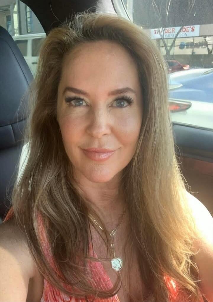 A woman takes a selfie in a car. In the background, a sign for 'Trader Joe's' is visible.