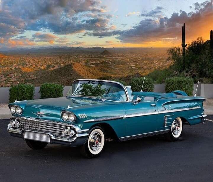 A classic turquoise convertible car parked in front of a desert landscape at sunset.