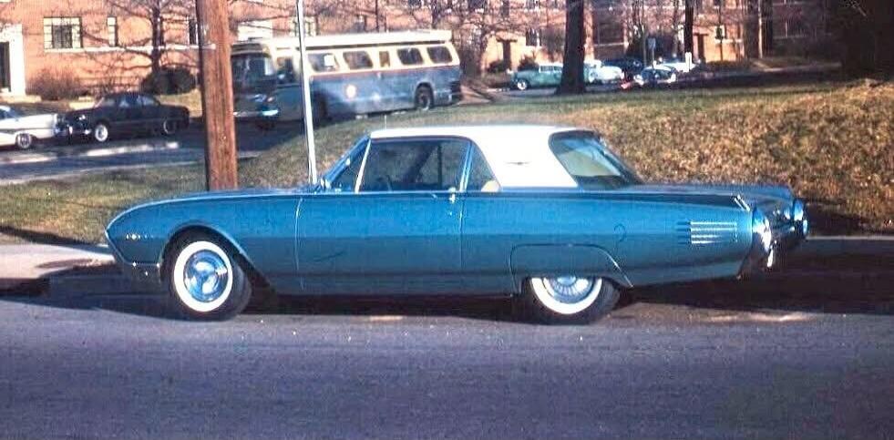 A vintage blue car with a white roof is parked on the side of the road. In the background, there is a bus and other cars. The scene appears to be from the mid-20th century.