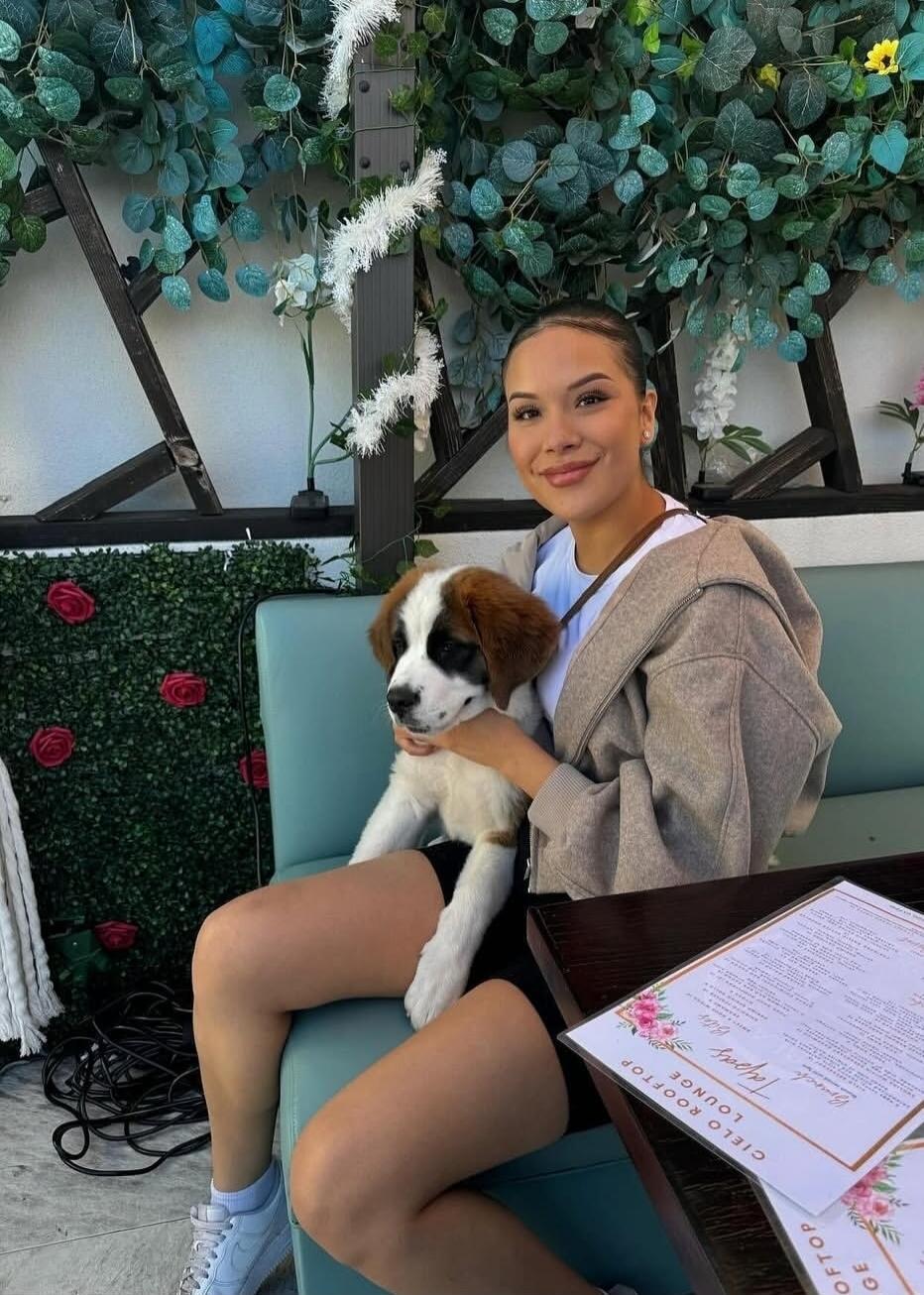 A woman sits on a light blue bench with a brown and white puppy on her lap, smiling. She is outdoors with greenery and flowers in the background.