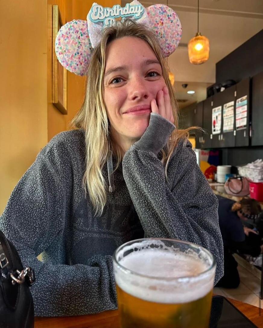 A young woman wearing a glittery 'Birthday' headband with mouse ears, sitting at a restaurant table with a beer in front, smiling with her hand on her cheek.