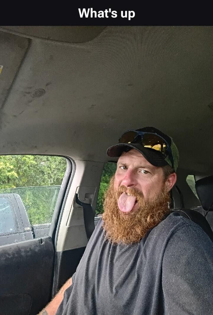 What's up. A man with a red beard is sitting in a car, sticking out his tongue and making a funny face.