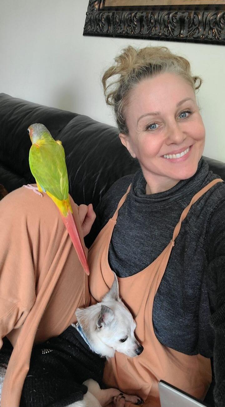 A woman with a parrot on her shoulder and a small white dog on her lap.