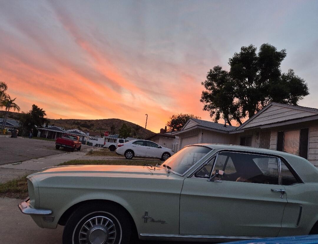 A classic car, possibly a Ford Mustang, is parked on a residential street. In the background, there are houses, hills, and a vibrant sunset sky with orange and pink clouds. A 'STOP' sign is visible.