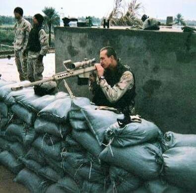 A soldier in camouflage uses a sniper rifle from behind a sandbag barrier.