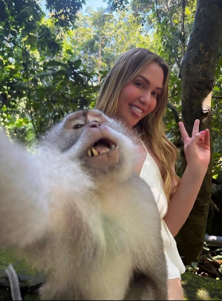 A woman smiling and making a peace sign while a monkey appears to be taking a selfie with a funny expression in a lush green jungle setting.
