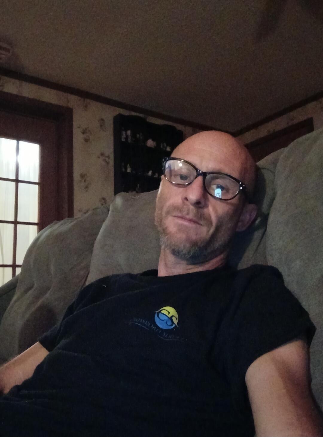 A man with glasses and a beard is sitting on a couch, looking directly at the camera. He is wearing a dark t-shirt with a logo. The room appears dimly lit.