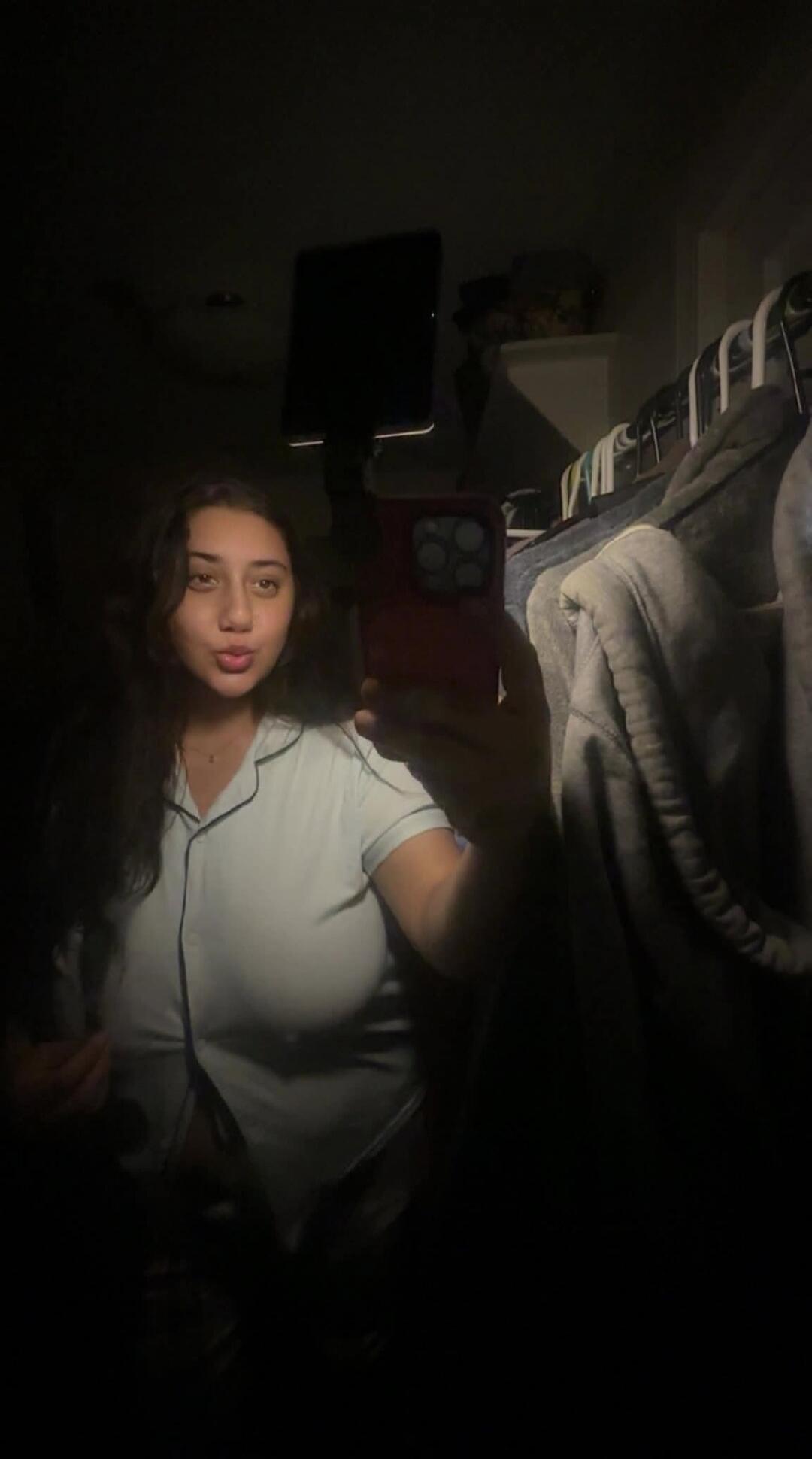 A young woman takes a mirror selfie in a dimly lit room. She is wearing a light blue pajama top, making a kissy face. Her long dark hair is down. In the background, a closet with clothes hangers is visible.