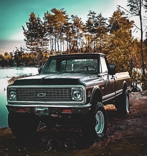A vintage Chevrolet pickup truck parked on a rocky lakeside trail with autumn trees in the background.
