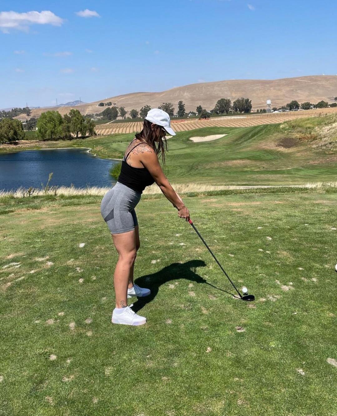 A woman in a black tank top, grey shorts, white cap, and white sneakers is playing golf on a sunny day. She is holding a golf club, preparing to hit the ball on a green golf course with a lake, hills, and trees in the background.