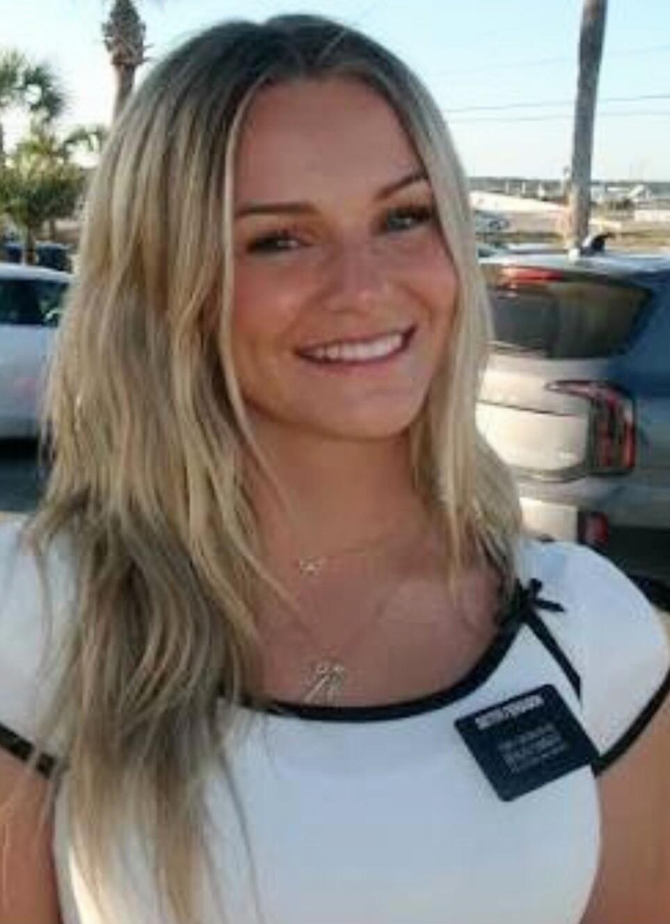 A young blonde woman is smiling at the camera. She is wearing a white top with black trim and a name tag that says 'SISTER' followed by some unreadable text, likely indicating she is a missionary. The background shows an outdoor setting with palm trees and cars.