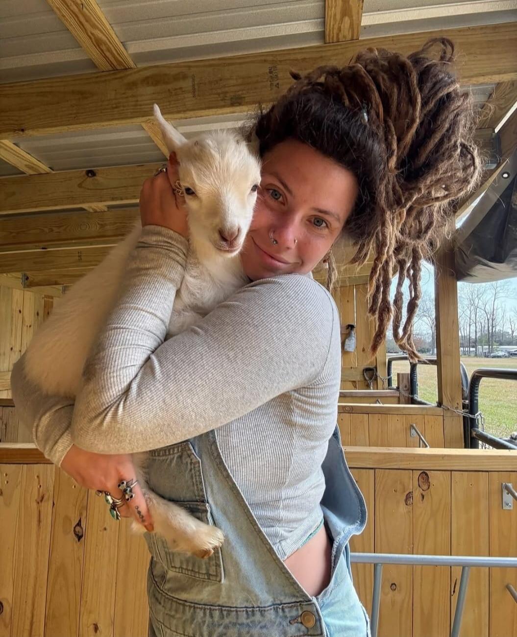 A person with dreadlocks is holding a small white goat and smiling at the camera in a barn.