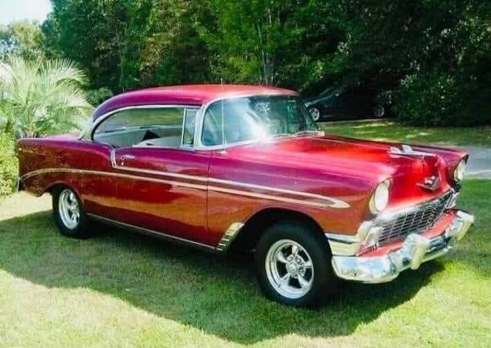 A classic red two-door car, possibly a Chevrolet Bel Air from the 1950s, is parked on a lush green lawn. The car features chrome accents, custom wheels, and a glossy paint finish. Trees and other foliage are visible in the background under natural light.