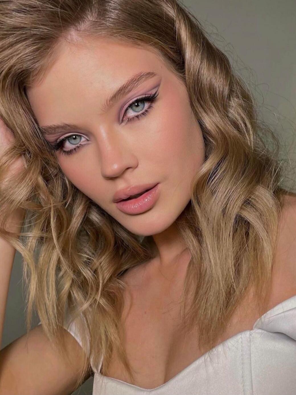 Portrait of a young woman with wavy blonde hair, winged eyeliner, pink lipstick, and a white top.