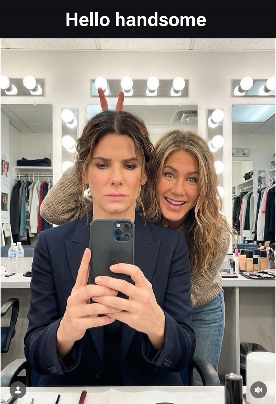 Hello handsome. Sandra Bullock takes a selfie with a serious expression while Jennifer Aniston playfully makes bunny ears behind her, smiling broadly in what appears to be a dressing room.