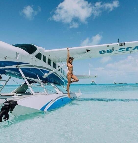 C6-SEA. A woman in a bikini poses on the float of a seaplane in clear turquoise water under a blue sky.